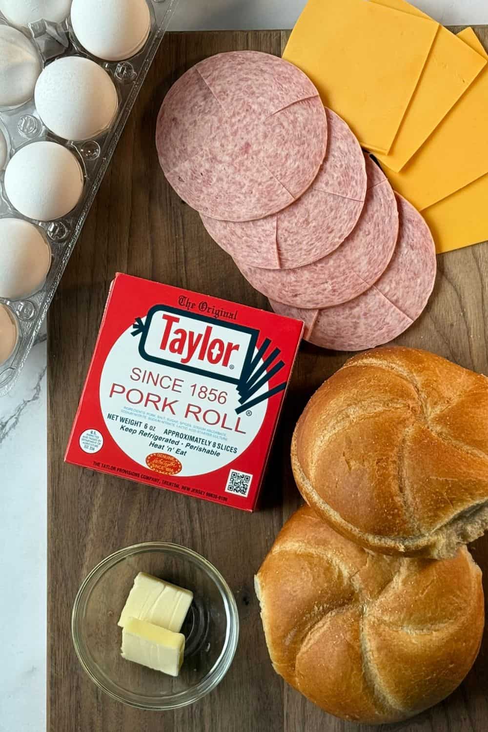 All the ingredients for a taylor ham, egg, and cheese breakfast sandwich laid out on a cutting board