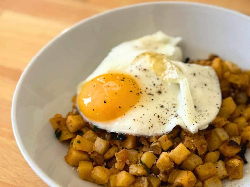 White bowl full of breakfast potatoes and topped with a fried egg