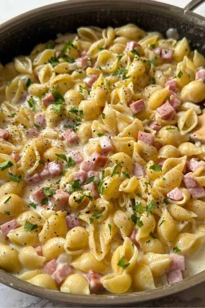 A skillet full of creamy ham and pasta and a wooden spoon