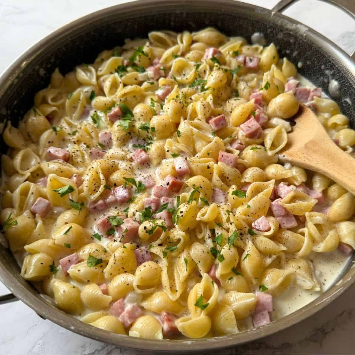 A skillet full of creamy ham and pasta and a wooden spoon