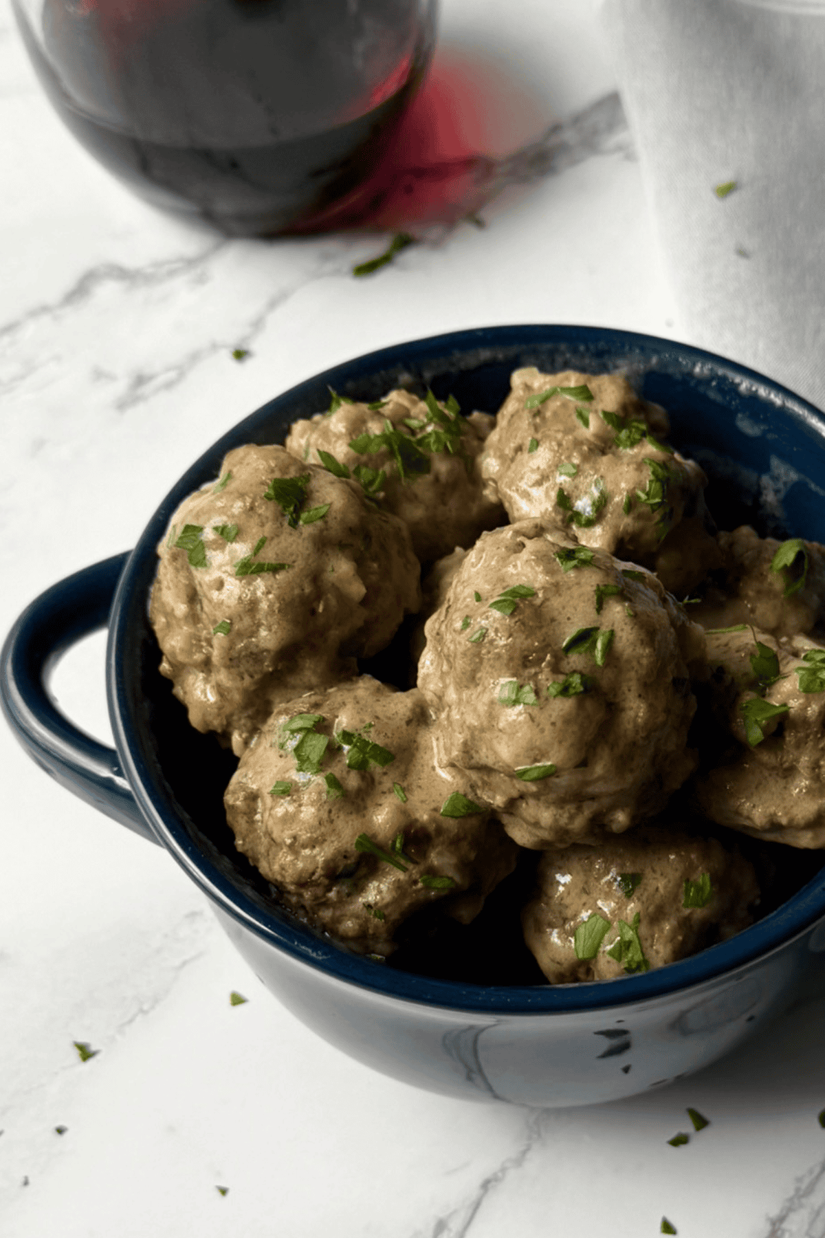 A blue bowl full of Swedish meatballs with a glass of red wine in the background
