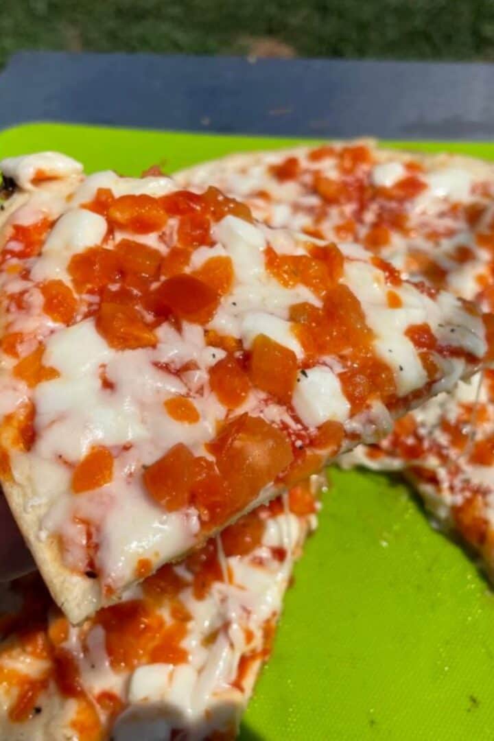 Frozen pizza cooked outside on a griddle on a green cutting board and a hand holding one of the slices