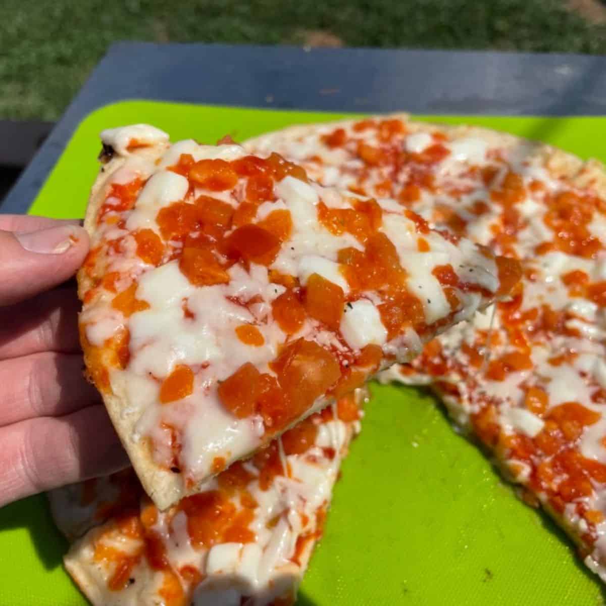 Frozen pizza cooked outside on a griddle on a green cutting board and a hand holding one of the slices