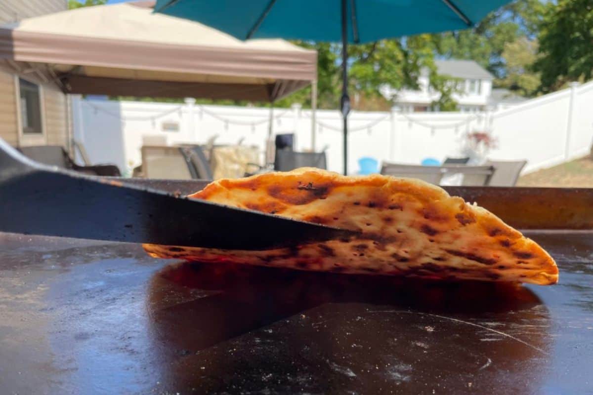 Frozen thin crust pizza cooking on a blackstone and being held up by a spatula to show the bottom browning