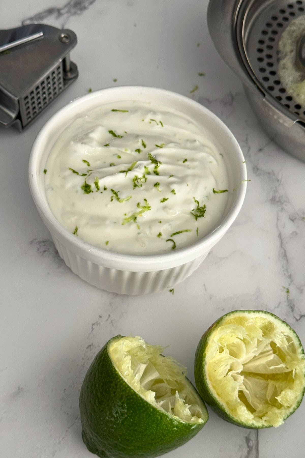 Bowl of garlic lime crema with some limes, a citrus squeezer and a garlic press