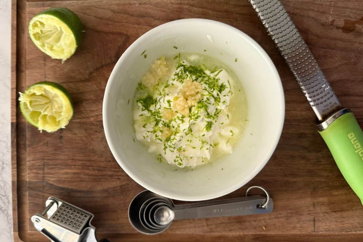 Bowl of garlic lime crema with some limes, a citrus squeezer and a garlic press