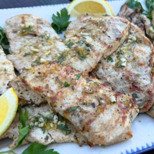 Slices of marinated grilled chicken cutlets on a platter and garnished with fresh parsley and lemon slices